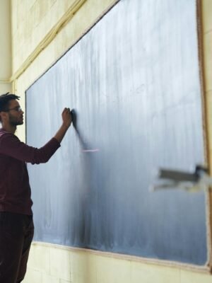 Man with dreadlocks writing on a blackboard in a classroom setting.